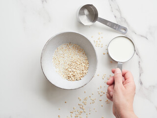 Hand holding half steel measuring cup with milk preparing oat porridge for breakfast in a bowl. Empty steel measuring cup on the table.
