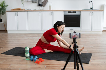 Woman in red activewear doing leg stretching exercise on yoga mat in modern kitchen. Filming fitness routine using smartphone. Healthy lifestyle, home workout, and active living concept.