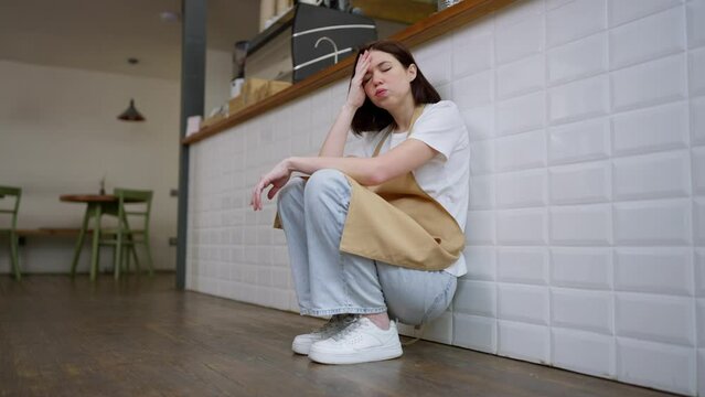 Tired girl brunette waitress sits near the cash register and rubs her forehead with her hands during a hard day at work in a cafe