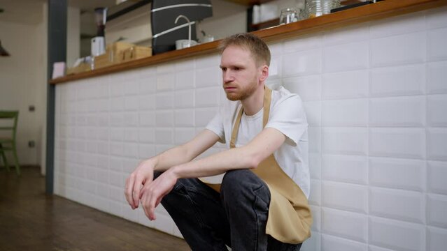 Tired cafe worker waiter sitting near a white wall and trying to take a break exhaling and massaging his head during a hard day at work