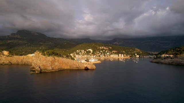 4K Aerial Drone video of popular summer destination, sunset over the cliffs with a lighthouse and a huge port of boats anchored at golden hour with seagulls flying. Port de Soller, Mallorca