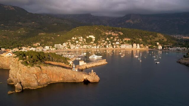 4K Aerial Drone video of popular summer destination, sunset over the cliffs with a lighthouse and a huge port of boats anchored at golden hour with seagulls flying. Port de Soller, Mallorca