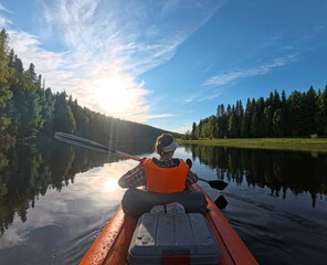 Kayaking Through Forested River in Summer Afternoon. The sun shines brightly in the blue sky, casting reflections on the water. The kayak is orange and the person is wearing an orange life vest