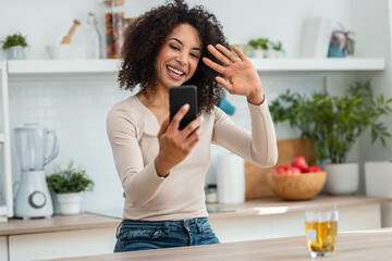 Beautiful woman doing a videocall with her smartphone while drinking a cup of tea in the kitchen at home.