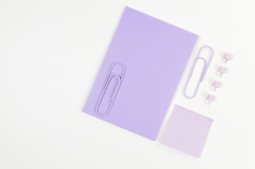 Top view of purple notebook on white background. Purple sticky note, paperclips and pins. School, office wallpaper. Flat lay, copy space.