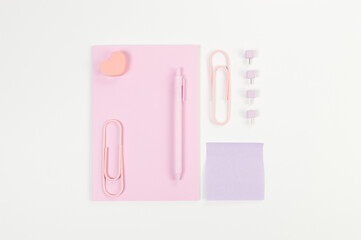 Top view of pink notebook on white background. Sticky note, paperclips, heart, pen and pins. School, office wallpaper. Flat lay, copy space.
