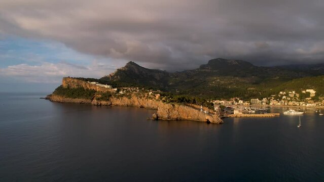 4K Aerial Drone video of popular summer destination, sunset over the cliffs with a lighthouse and a huge port of boats anchored at golden hour with seagulls flying. Port de Soller, Mallorca