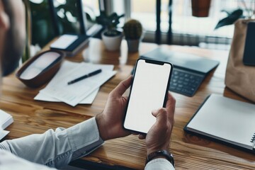 Mockup of a man's hands holding smart phone with blank white screen while sitting at the wooden table in modern office