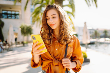 Fototapeta premium Young beautiful woman walking along the street of the city and uses a phone, smiling, feel happy. Technology, blogging, communication concept.