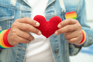 Asian lady wearing rainbow flag wristbands and hold red heart, symbol of LGBT pride month celebrate annual in June social of gay, lesbian, bisexual, transgender, human rights.