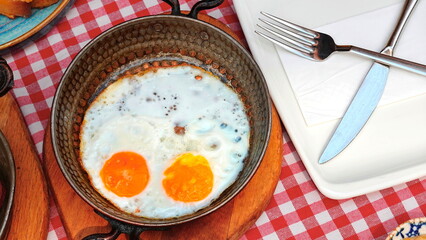 wo perfectly fried eggs sizzle in a skillet, set on a table with a charming red checkered tablecloth. A plate with a fork and knife awaits, ready to indulge in this delightful breakfast delight