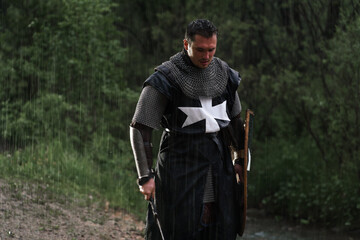 Crusader knight in armor with sword in rain in forest