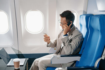 Young Asian executive excels in first class, multitasking with digital tablet, laptop and...