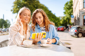 Two young women, sisters, best friends showing news social media photos on smart phone cellphone sitting in cafe and drinking coffee during breakfast. Fashion, beauty, blogging, tourism.