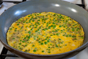 Delicious omelette cooking in frying pan, mixture of eggs, garden peas, herbs. Selective focus.