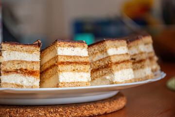 Honey semolina cream of a layered tea cake slice with apricot jam in the middle and chocolate coating on top, aesthetically arranged in a line on a white ceramic plate and wooden table.