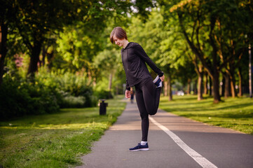 Obraz premium A young woman in black sportswear stretching her leg on a park path before running. Highlighting warm-up routines and a healthy outdoor lifestyle.