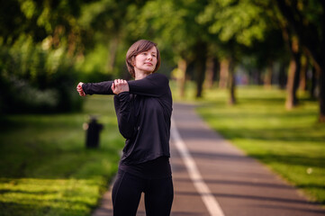 A young woman in black athletic wear stretching her arm on a park path before running. Emphasizing...