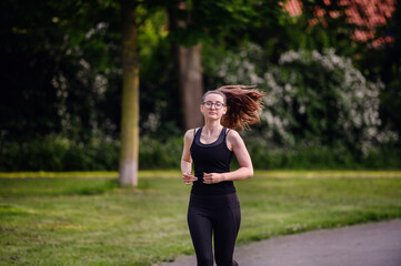 A young woman in black sportswear running on a park path, with her hair flowing in the wind....