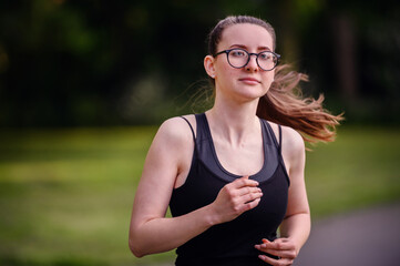 A young woman in black sportswear and glasses running in a park, showcasing determination and a...