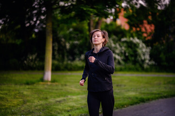 A young woman in black sportswear jogging in a park at dusk, showcasing determination and a healthy lifestyle.