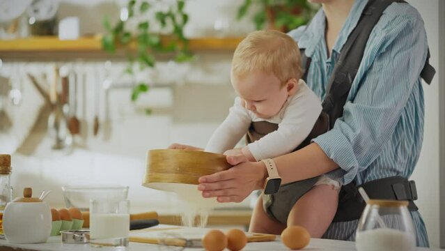 A housewife uses a sling for a child while cooking. The child touches the flour with his hands in the kitchen. Modern woman methods of raising children uses the latest sling for convenience, ergonomic
