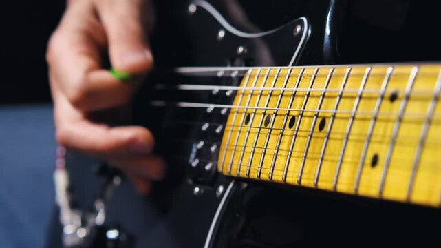 Close-up of a guitarist's hand playing a black electric guitar, with emphasis on the fret and strings. Fingers holding a green plectrum
