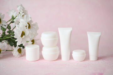 White tubes and jars of cream with bouquet of flowers on a pink background