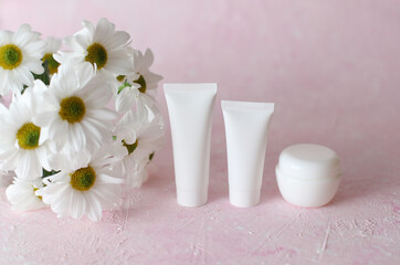 White tubes and jars of cream with bouquet of flowers on a pink background