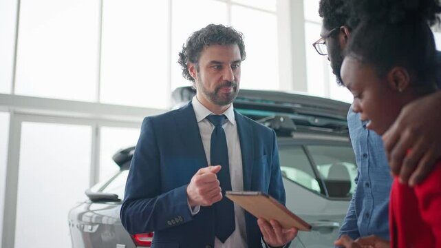 Manager explaining something to clients in auto centre. Married couple with enthusiasm looking at different models of cars on tablet device. People watching at vivid images and videos in presentation.