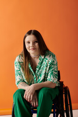 A smiling woman in a wheelchair, wearing a green floral outfit, sits against a vibrant orange backdrop confidently.