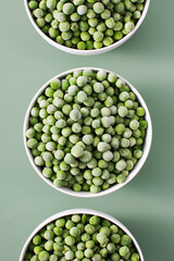 Three white bowls filled with frozen green peas on green background.