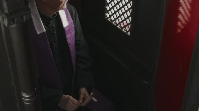 Tilt up shot of senior priest in black robe and purple stole sitting inside of confession booth and looking through lattice screen