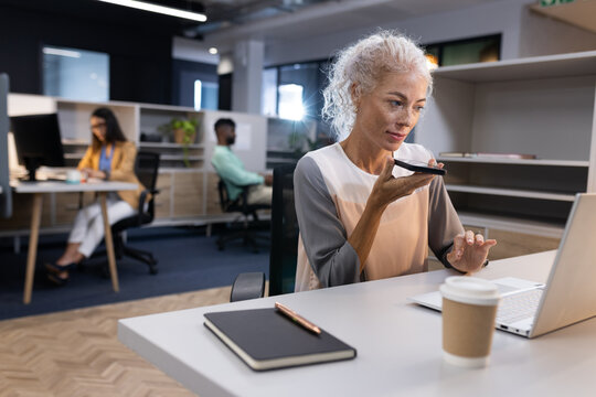 Using voice command on smartphone, woman working on laptop in modern office