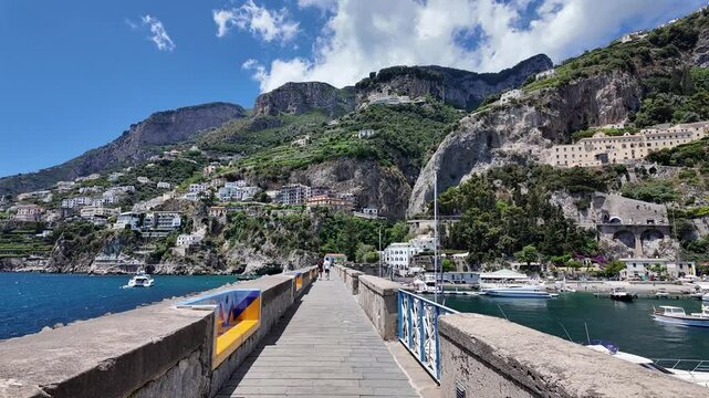Walk on the pier at Marina Coppola in Amalfi Harbor, surrounded by the Tyrrhenian Sea, the Gulf of Salerno, majestic mountains, lush vegetation, and boats in Italy