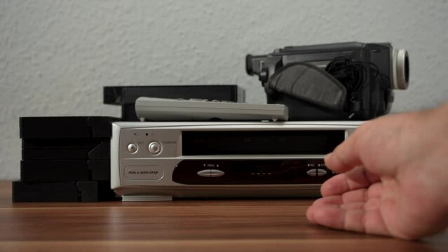 A small silver VHS player sits on a wooden table next to a stack of black cassette tapes. Silver and black VCR.