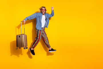 Full size photo of pretty young guy suitcase traveler running winning wear trendy jeans outfit isolated on yellow color background
