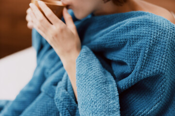 Woman wrapped in blue blanket sitting on bed in bedroom with cup of coffee in hand
