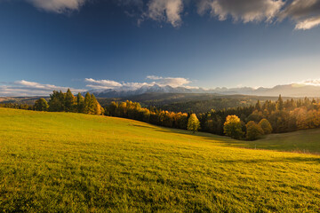 A beautiful autumn landscape of the Tatra Mountains seen from Polish Podhale during the golden hour. The sky is painted in warm hues, with the mountains bathed in golden light.