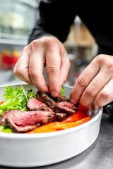 A chef’s hands garnishing a gourmet dish with greens, featuring medium-rare steak and fresh vegetables.