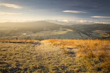 A beautiful autumn landscape of the Tatra Mountains seen from Polish Podhale during the golden hour. The sky is painted in warm hues, with the mountains bathed in golden light.