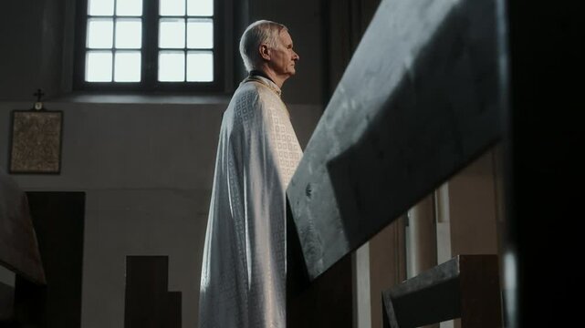 Side view of senior pastor dressed in white chasuble walking among empty pews in Lutheran church