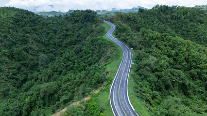 Aerial view from above of country road through the green summer forest in summer Thailand. Top view of the asphalt road and dense green forests.