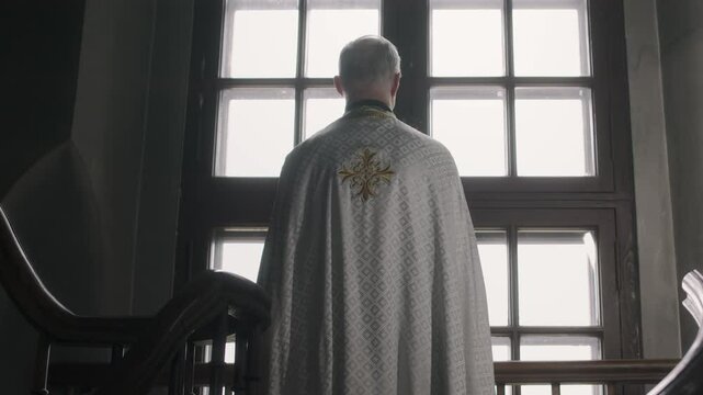 Tilt up back view shot of pastor in traditional chasuble standing on spiral staircase in church and looking through window