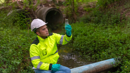 An ecologist in a green jacket sitting on a blue pipe while holding the glass tube on his left hand...