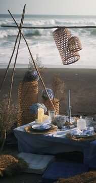 Picnic on black sand with big ocean waves in the background. The table is decorated with a blue tablecloth and dishes, there are candles and flowers on it. Summer holidays.