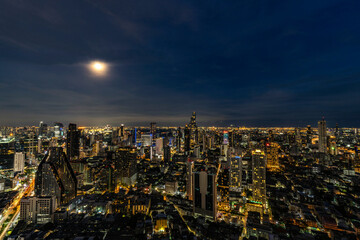 A breathtaking night view of Bangkok's skyline illuminated by city lights and a full moon. The cityscape showcases the blend of modern skyscrapers and vibrant urban life, reflecting the city's dynamic