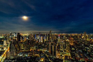 Obraz premium A breathtaking night view of Bangkok's skyline illuminated by city lights and a full moon. The cityscape showcases the blend of modern skyscrapers and vibrant urban life, reflecting the city's dynamic