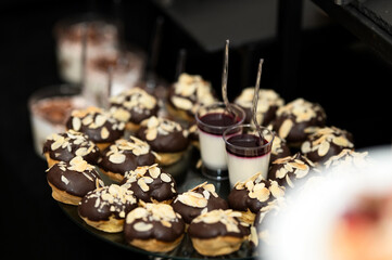 Small Eclairs in chocolate glaze. Festive Dessert on plate. Profiteroles are decorated with almond petals. Mousse, jelly in glass with spoon. Sweet buffet. Side view. Dark background. Soft focus