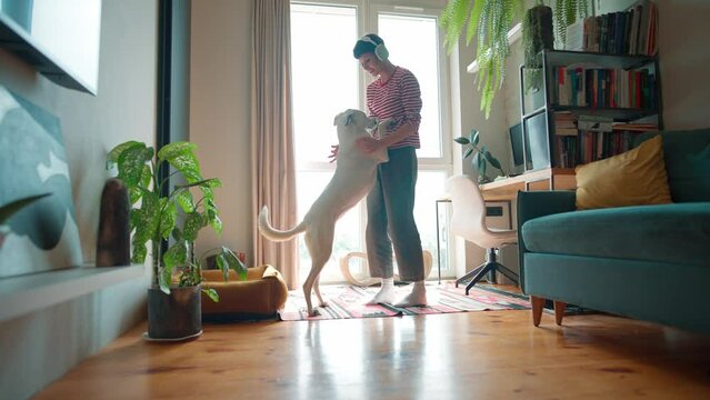 Side view of Caucasian woman wearing headphones. Pretty female holding paws of her dog while standing before large panoramic window. Happy owner dancing with her lovely pet in cosy home.